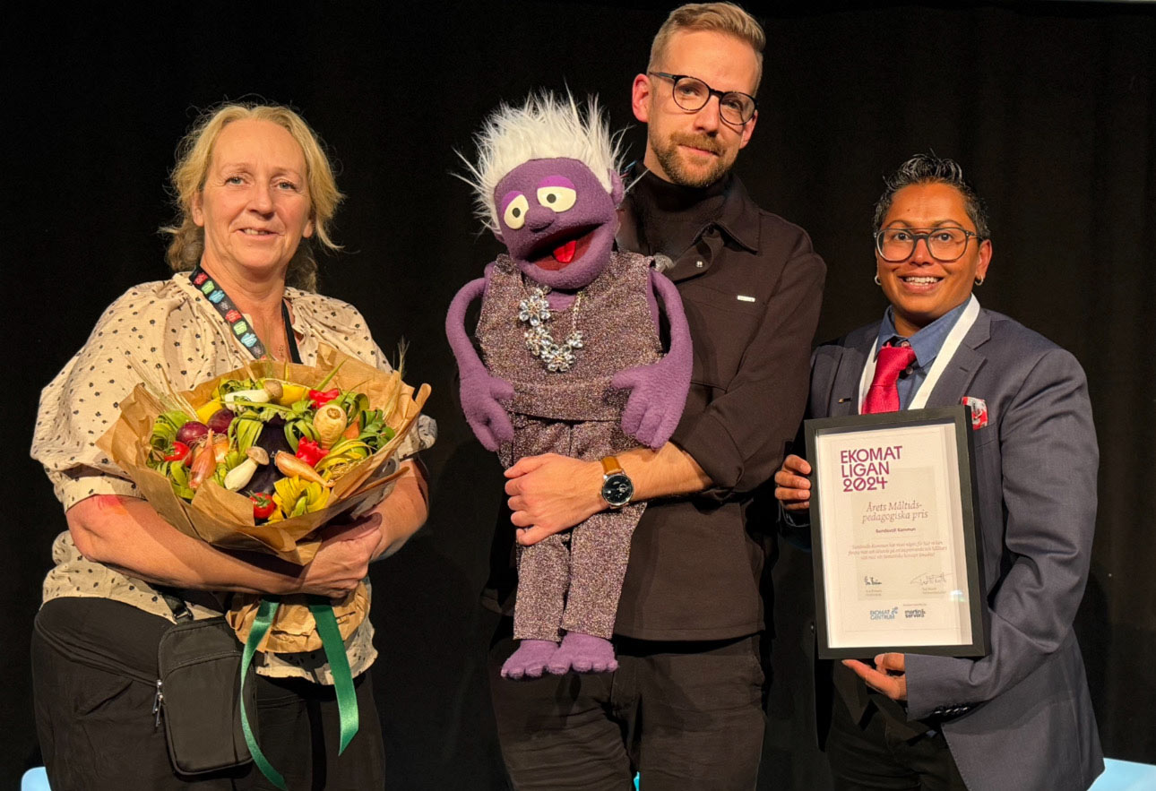 Tre personer står tillsammans med den lila tygdockan Smaskis på en scen, med diplom och blommor