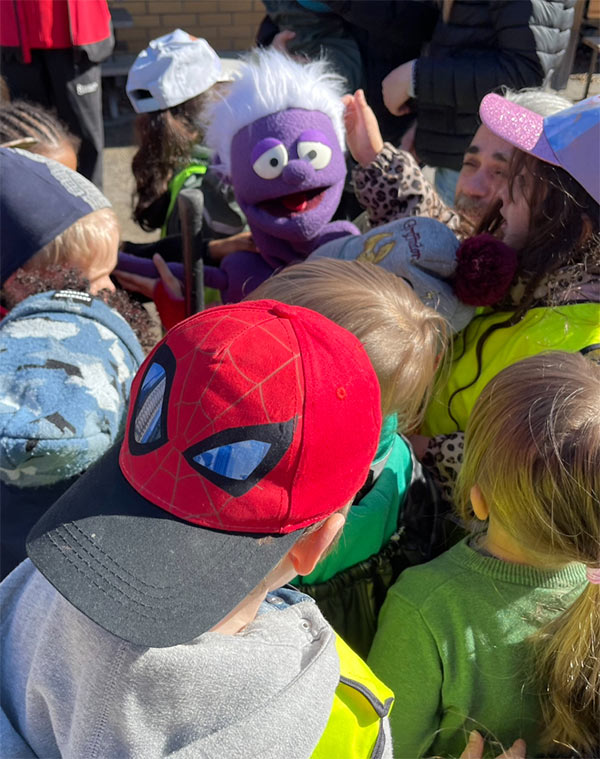 Den lila tygdockan Smaskoten Smaskis omringad av glada barn på en skolgård