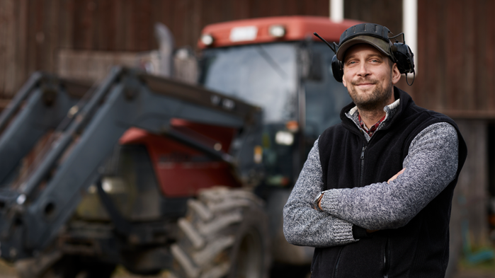 Ung lantbrukare står bredvid en röd traktor framför en maskinhall. Personen har grå tröja och hörselkåpor på huvudet