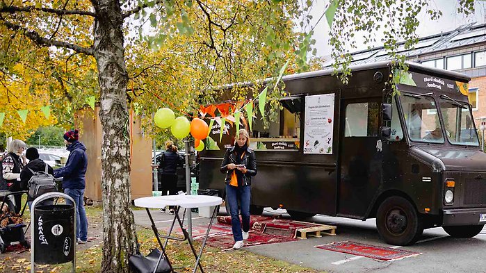 En foodtruck som står uppställd i en park. I förgrunden syns invånare som provsmakar mat.